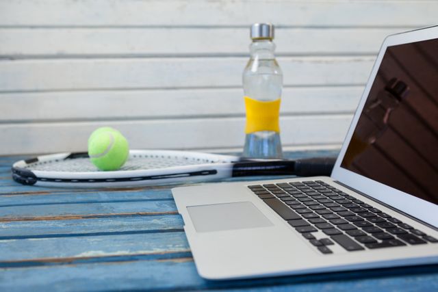 Close up of laptop by tennis racket and balls with water bottle on wooden table against white wall