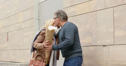 Mature couple sharing kiss on city sidewalk while woman holding bouquet and leather bag