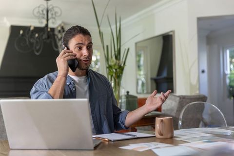Remote professional talking on smartphone while working at home desk with laptop and notes