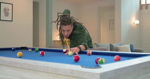 African american man focusing while playing pool on blue-felt table in modern living room