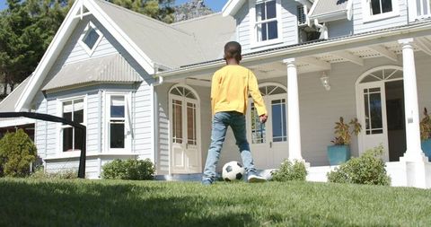 Child playing soccer on green lawn in front of classic white suburban home with porch