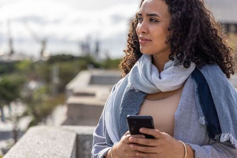 Woman standing on stone balustrade holding smartphone and gazing toward harbor skyline