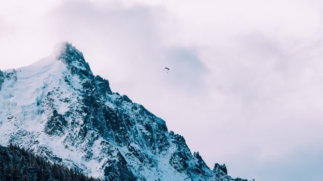 Stunning view of a snow-covered mountain peak with a lone paraglider in the cloudy sky. Ideal for advertisements and editorials focused on adventure sports, winter activities, nature exploration, and travel promotions. Can be used in websites, magazines, brochures, or any other visual content relating to the beauty and thrill of outdoor adventures in alpine regions.