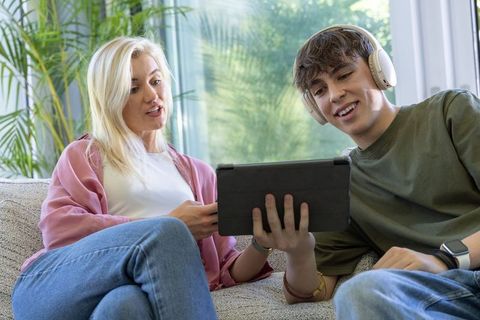 Mother and teenage son sharing tablet and headphones on sofa relaxing at home
