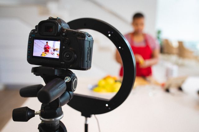 African American female food blogger recording a cooking video in a modern kitchen. She is using a digital camera mounted on a tripod with a ring light. This image is perfect for illustrating concepts related to food blogging, content creation, social media influencers, culinary arts, and video production. Ideal for use in articles, blogs, and marketing materials focused on digital content creation, cooking tutorials, and influencer marketing.