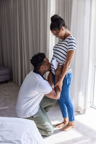 Kneeling partner kissing pregnant belly while partner holding love letter, tender moment