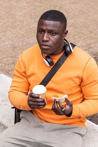 African american man sitting wearing orange sweater holding coffee and muffin with headphones