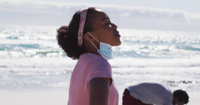 African American woman on sunny beach wearing a face mask symbolizes a blend of health precautions and enjoyment during the pandemic. This can be used in materials related to travel during health crises, promoting safety while enjoying outdoor activities, and pandemic lifestyle changes.