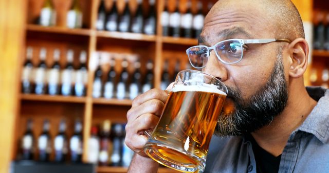 Middle-aged man enjoying a refreshing beer in a bar setting, surrounded by shelves of bottled beverages. Perfect for use in content about relaxation, socializing, nightlife, bars, and alcoholic beverages. Ideal for advertisements, blog posts, or articles on beer culture, craft breweries, and pub experiences.