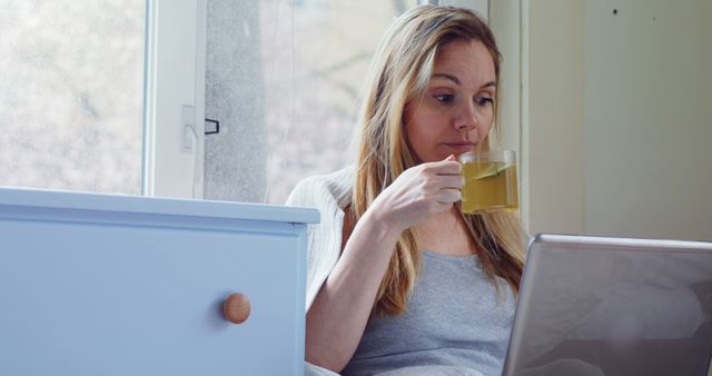 Woman sitting on bed or sofa by window while drinking herbal tea and using laptop. Ideal for articles on remote work, relaxation at home, healthy living, casual lifestyle, or morning routines. Image evokes feelings of comfort, tranquility, and productivity in a homey setting.