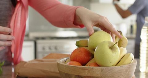 Woman reaching for green apple in wooden fruit bowl on kitchen counter, couple morning