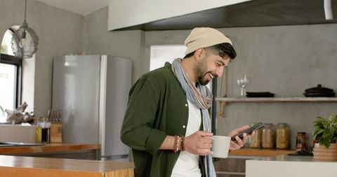 Indian young man checking smartphone and holding coffee mug in modern kitchen, smiling