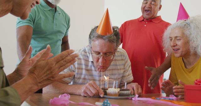 Senior man wearing orange party hat blowing out birthday candles on cupcake surrounded by cheering friends. Ideal for use in retirement community promotions, birthday party advertisements, and content highlighting active elderly social lives.