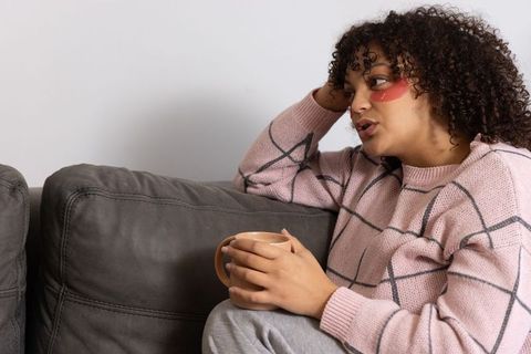 Relaxed woman wearing under-eye patches holding mug on couch enjoying cozy self-care moment