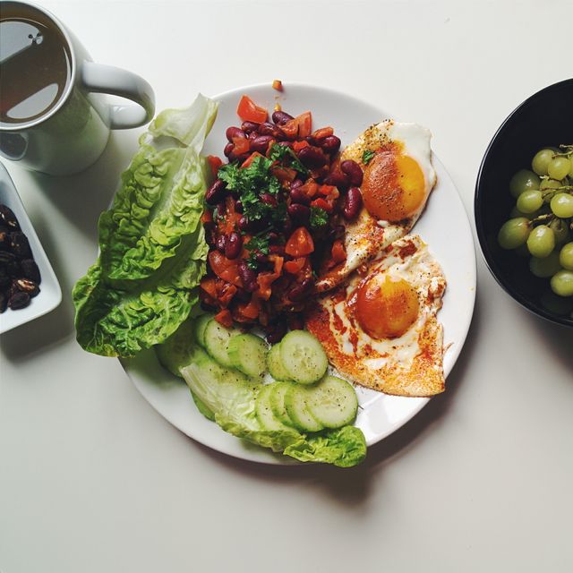 Delicious and nutritious breakfast featuring fried eggs, kidney beans with diced tomatoes, lettuce, cucumber, black coffee, and green grapes. This image is ideal for use in articles about healthy eating, balanced diets, nutritious recipes, meal planning, and morning routines.