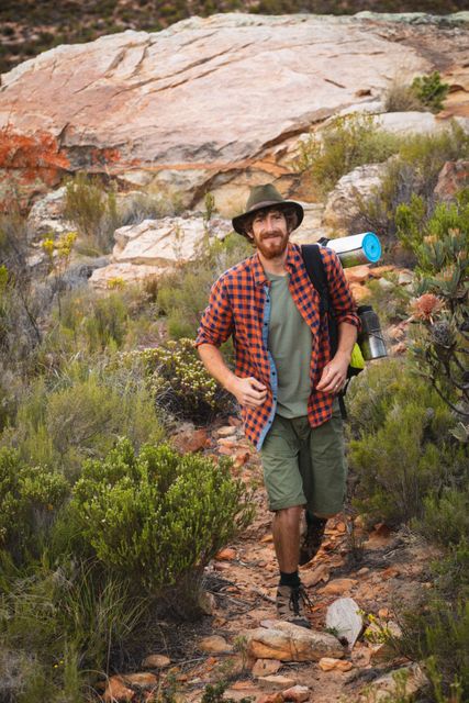 Smiling male hiker walking on a rocky trail surrounded by plants, carrying a backpack and wearing a hat. Ideal for use in content related to outdoor adventures, hiking, survivalism, weekend activities, and nature exploration.