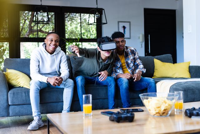 Teenage friends are sitting on a couch in a living room, with one wearing a VR headset and playing a game while the others watch and smile. This image can be used to depict themes of friendship, modern technology, and leisure activities. Ideal for use in advertisements for gaming products, social media posts about youth culture, or articles on the impact of technology on social interactions.