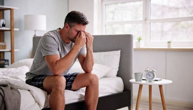 Tired middle-aged man rubbing eyes while sitting on bed in morning light with alarm clock