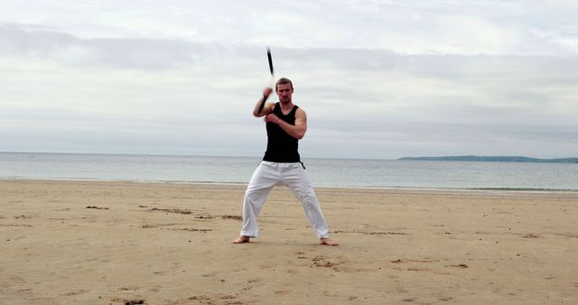 A man is practicing martial arts techniques with a weapon on a sandy beach. The man, dressed in a black tank top and white pants, shows concentration and strength during the exercise. This image is ideal for content related to martial arts training, physical fitness, outdoor workouts, and mindfulness activities by the sea.
