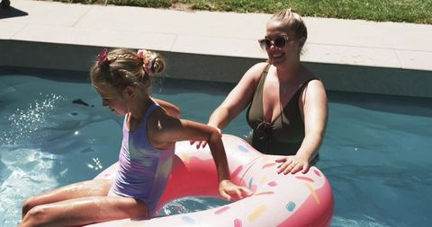Mother steadying daughter on pink sprinkles donut float in sunlit backyard pool