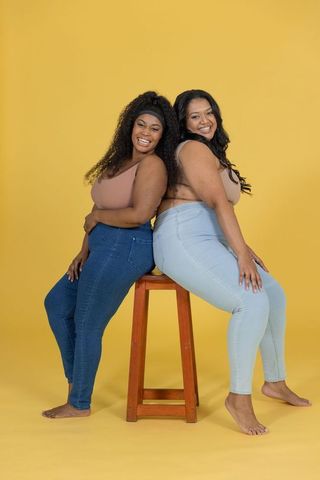 Smiling diverse plus-size women leaning back-to-back on stool wearing denim and cropped tanks
