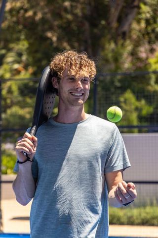 Curly-haired man tossing tennis ball on paddle court holding paddle over shoulder