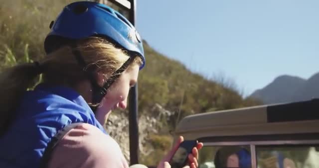 Young woman sits wearing protective gear including a blue helmet, using a smartphone while seated in an off-road vehicle. Sun shines brightly in a mountainous landscape background. Perfect representation for travel blogs, adventure vacation brochures, and outdoor activity promotions.