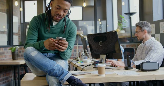 Person is seated casually on a desk looking at a smartphone, while a coworker is working on a computer in a bright, modern office environment. It can be used for websites, articles, and promotional materials related to modern workspaces, remote work, technology in the workplace, and teamwork.