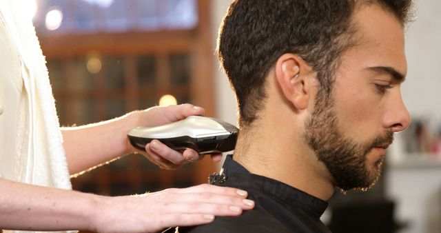 A young Caucasian man is getting a haircut from a hairstylist using electric clippers, with copy space. Capturing a moment of grooming, the image reflects personal care and the profession of hairstyling.