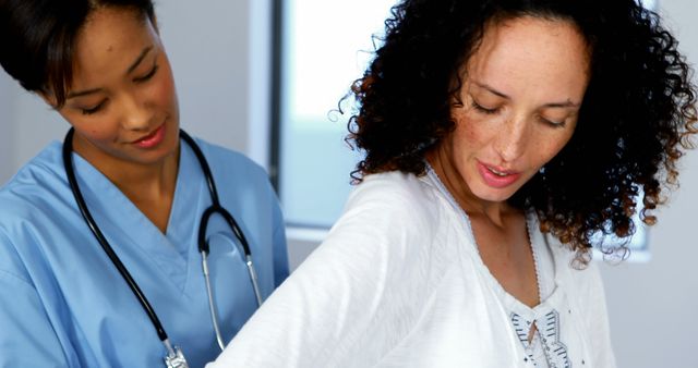 Image depicts a female nurse examining a patient at a medical appointment. Nurse wearing a blue scrub uniform with a stethoscope around neck assists patient, who appears to be showing her back. Suitable for healthcare-related articles, medical brochures, online health resources, and wellness promotion campaigns.