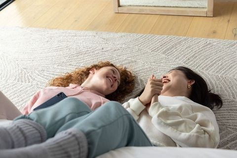 Sunlit cozy moment of diverse female friends lying on rug, holding hands and laughing