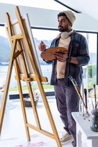 Male artist painting on easel in bright lakeside studio wearing headphones and palette