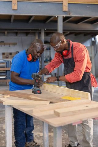 African american carpenters measuring wood and prepping cordless drill at workshop bench
