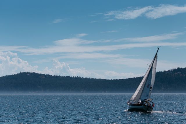 Sailboat gliding gently on a calm blue ocean under a bright and partly cloudy sky. The serene water and picturesque backdrop of distant forested mountains make it ideal for themes of travel, adventure, relaxation, and maritime activities. Perfect for use in travel brochures, adventure gear advertisements, or relaxation-themed publications.