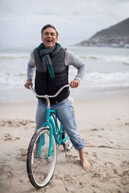 This image depicts a mature man enjoying a leisurely ride on a bicycle along a sandy beach. He is dressed in casual clothing with a scarf, suggesting a cool day by the ocean. The man is smiling, conveying happiness and a carefree attitude. This image can be used for promoting active lifestyles, vacation destinations, or products related to outdoor activities and leisure.