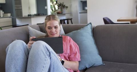 Young woman relaxing on sofa using tablet in bright modern living room with kitchen