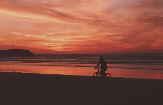 Silhouette of cyclist riding along a beach during sunset, featuring rich orange and red sky hues, adding a serene and peaceful atmosphere. Ideal for use in travel promotions, relaxation or leisure themes, lifestyle blogs, and beach-related activities ads.