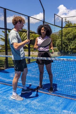 Mixed-race players chatting at net on blue padel court holding paddles and ball under sun