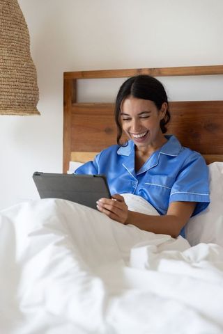Smiling woman in blue satin pajamas using tablet while relaxing in cozy white bed