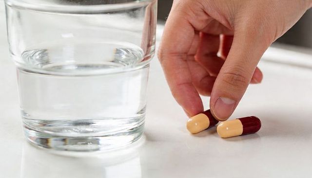 Adult hand picking up beige maroon capsules next to glass of water on white countertop