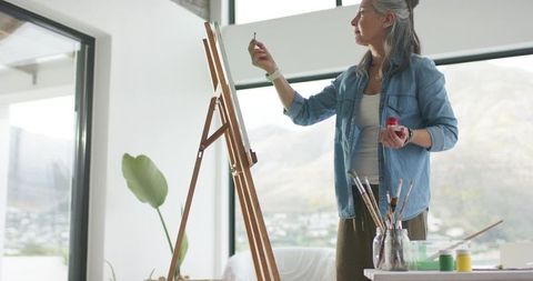 Senior woman painting on easel in bright minimalist studio with mountain view