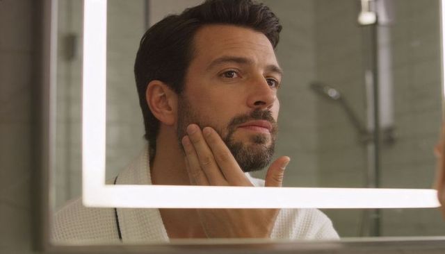 Man inspecting beard while grooming in led mirror wearing waffle robe in modern bathroom