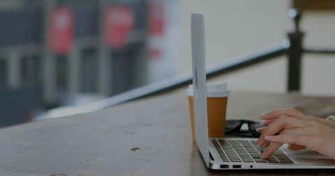 Freelancer typing on laptop at outdoor table with coffee and sunglasses, terrace workspace