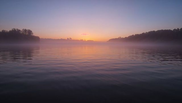 Reflecting sunrise mist on calm lake with pastel sky, ripples and woodland silhouettes