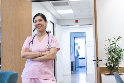 Smiling female nurse standing in clinic lobby wearing pink scrubs, stethoscope, smartwatch