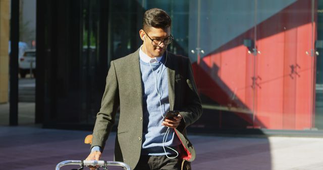 Young businessman walking in a modern urban area carrying a bicycle and looking at his smartphone. He is wearing casual yet professional attire, including eyeglasses and earphones. This image can be used for themes related to business, technology, urban lifestyle, commuting, and transportation.