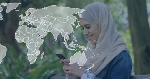 Woman wearing headscarf scrolling smartphone on park bench with world map overlay