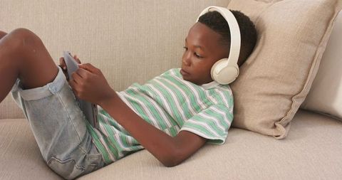 Relaxing young boy listening with headphones and using tablet on beige sofa at home