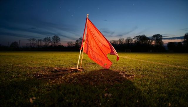Red corner flag fluttering on worn soccer pitch at dusk evoking matchday nostalgia