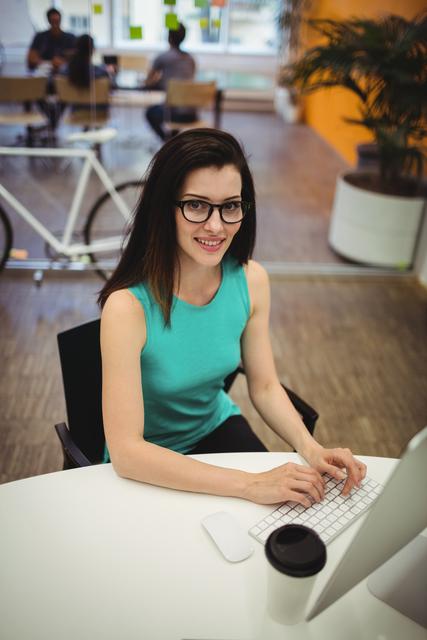 Young female executive smiling while working at her desk in a modern office. She is wearing glasses and casual attire, with a coffee cup on the desk. Ideal for use in business, corporate, and technology-related content, showcasing a professional and productive work environment.